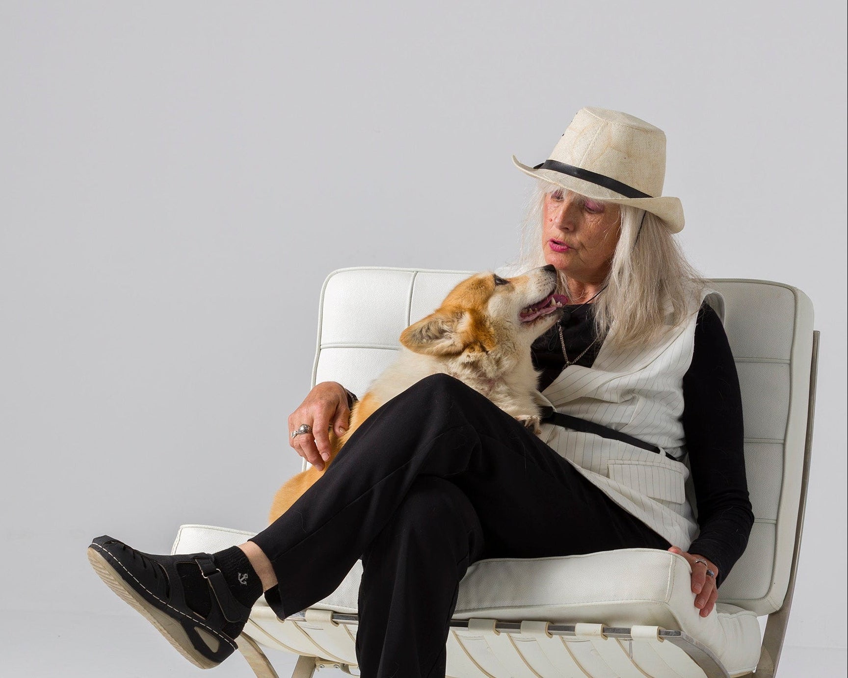 A woman wearing a cream fedora, black outfit, and white vest sits in a white leather lounge chair with a corgi on her lap, set against a seamless light gray studio background.